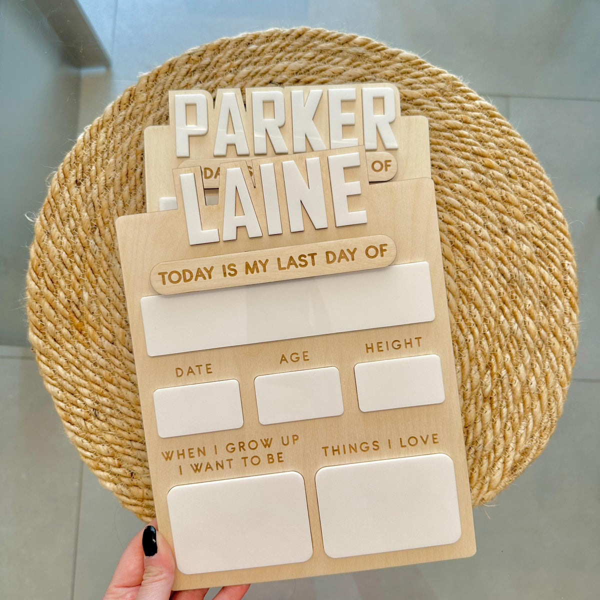 A wooden personalized milestone plaque held by a hand, featuring the text 'PARKER LAINE' and sections for date, age, height, and reflection prompts. The plaque is styled on a round woven jute placemat over a light gray tiled floor.

